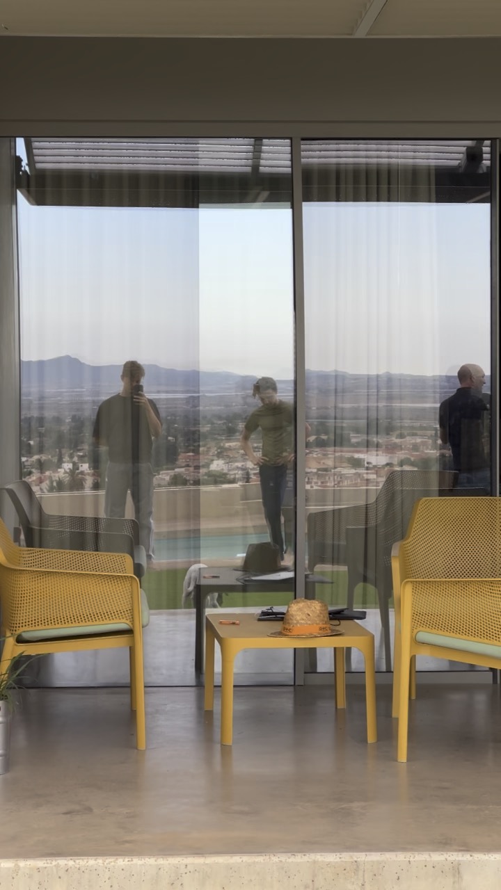 three people reflected in a glass door, with yellow balcony chairs and a mountain view